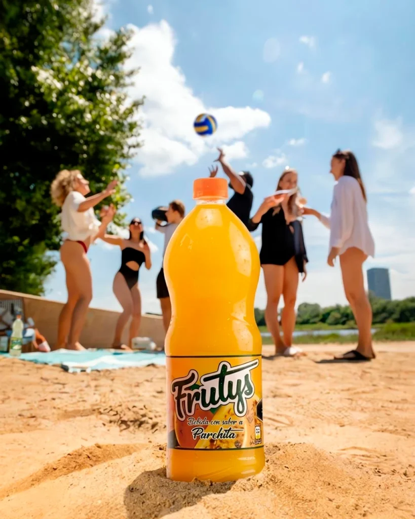 Botella de bebida Frutys sabor a parchita sobre la arena en un día de playa con amigos jugando voleibol.