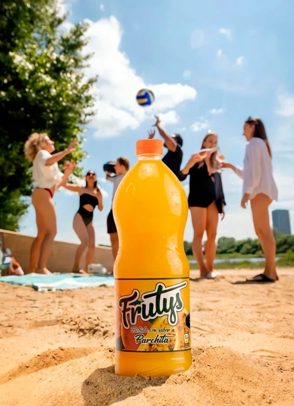 Botella de bebida Frutys sabor a parchita sobre la arena en un día de playa con amigos jugando voleibol.